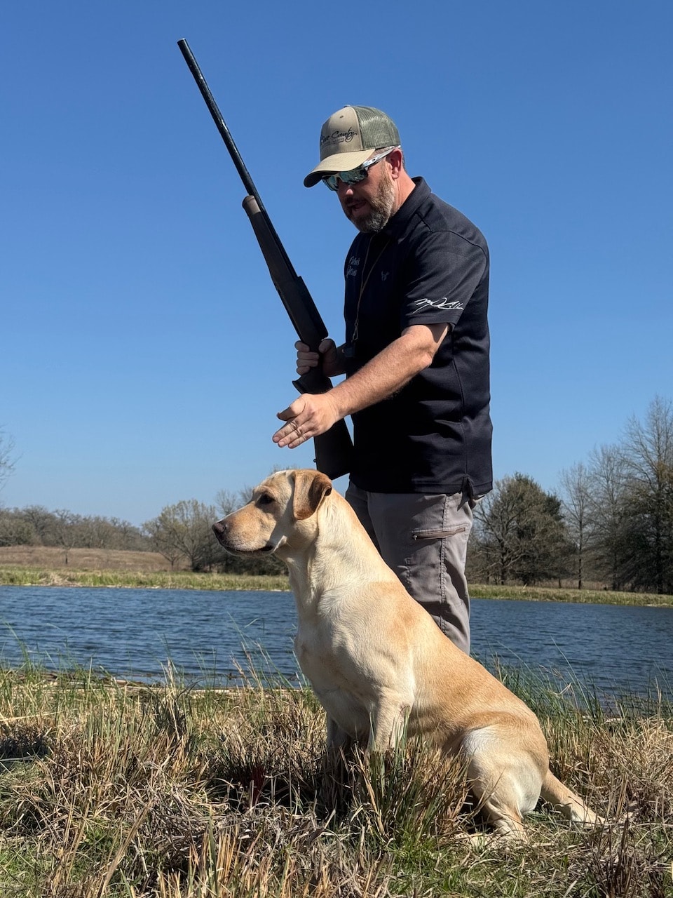 Trainer with yellow Labrador steady at heel beside pond