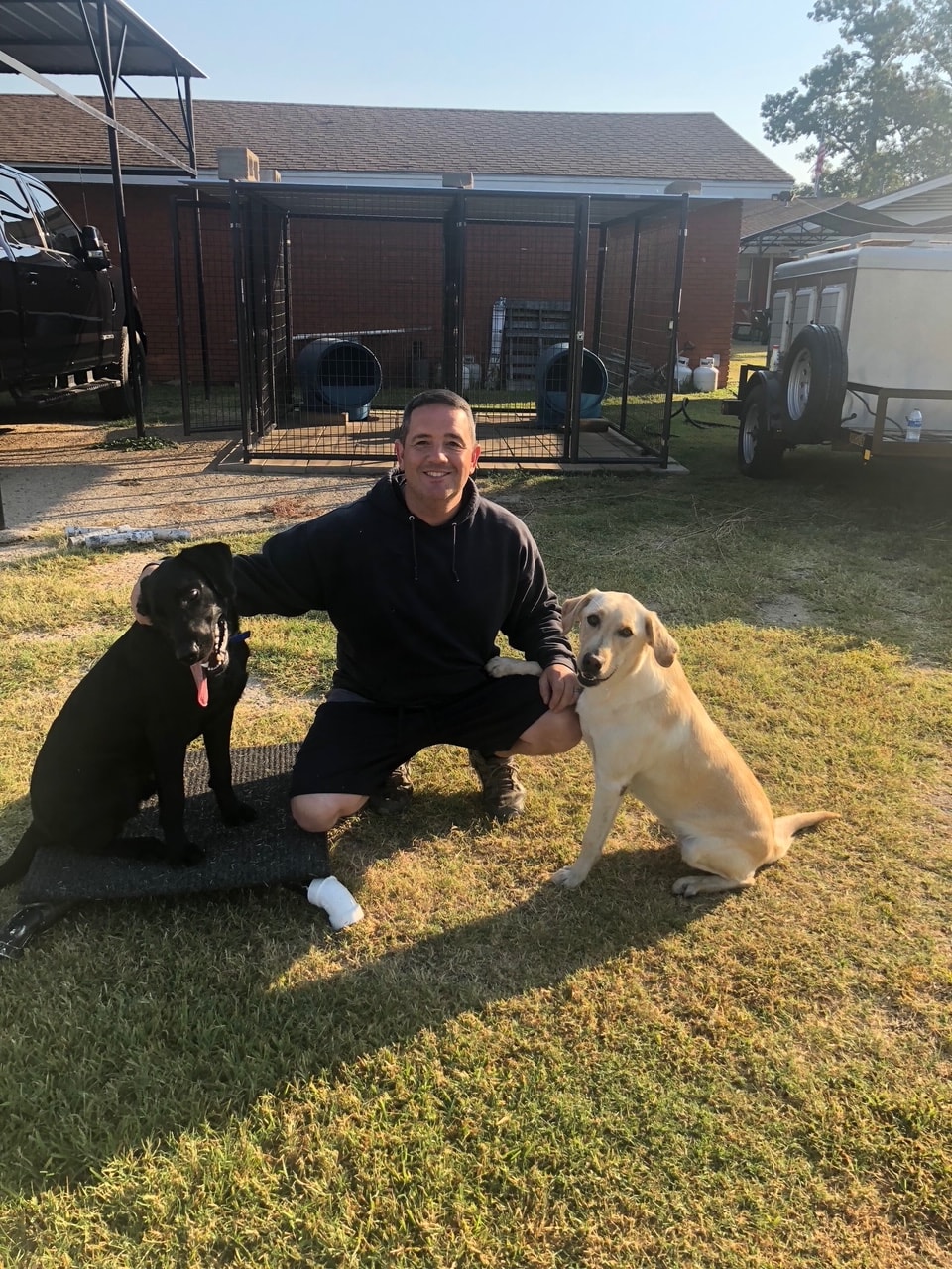 Trainer kneeling with black and yellow Labrador in backyard kennel